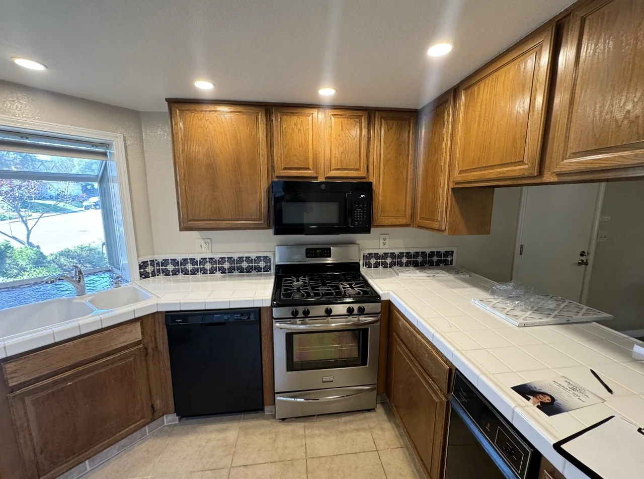 Kitchen before remodeling with oak cabinets and tiled counters beside a corner window